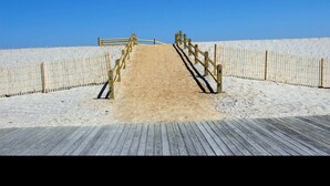 Ein hölzerner Steg führt zu einem Sandstrand, beidseitig von einer Holzlattenumrandung gesäumt.