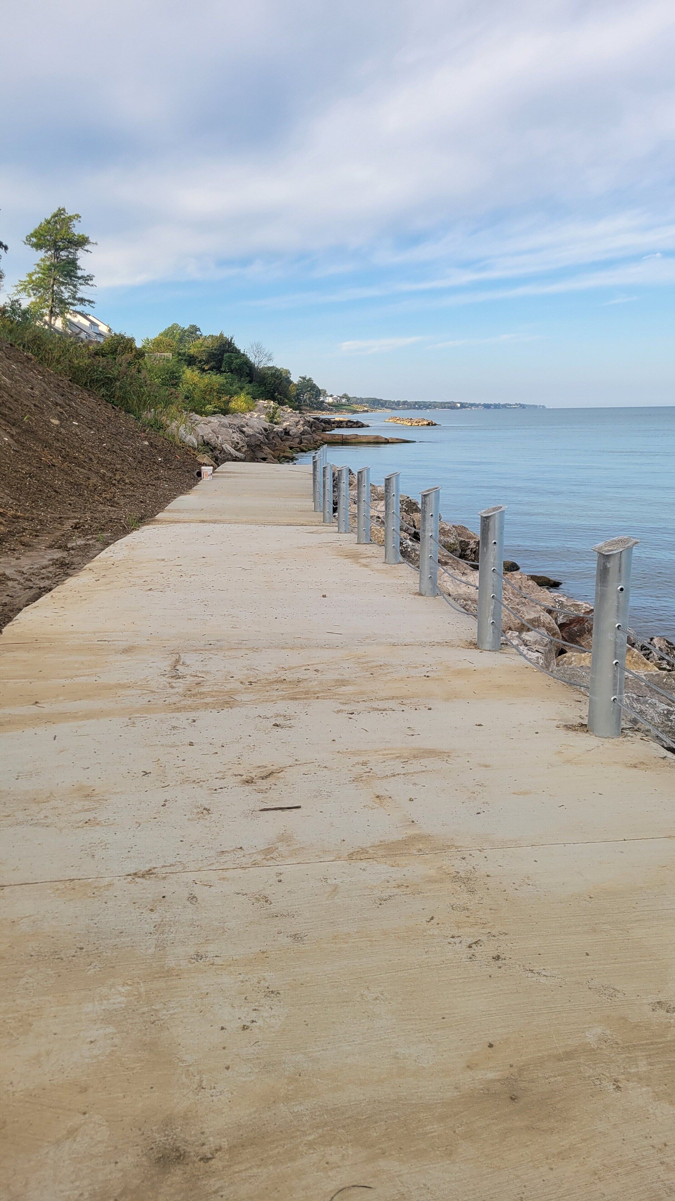 Un camino de hormigón bordea una costa rocosa con postes metálicos y una vista despejada del mar.