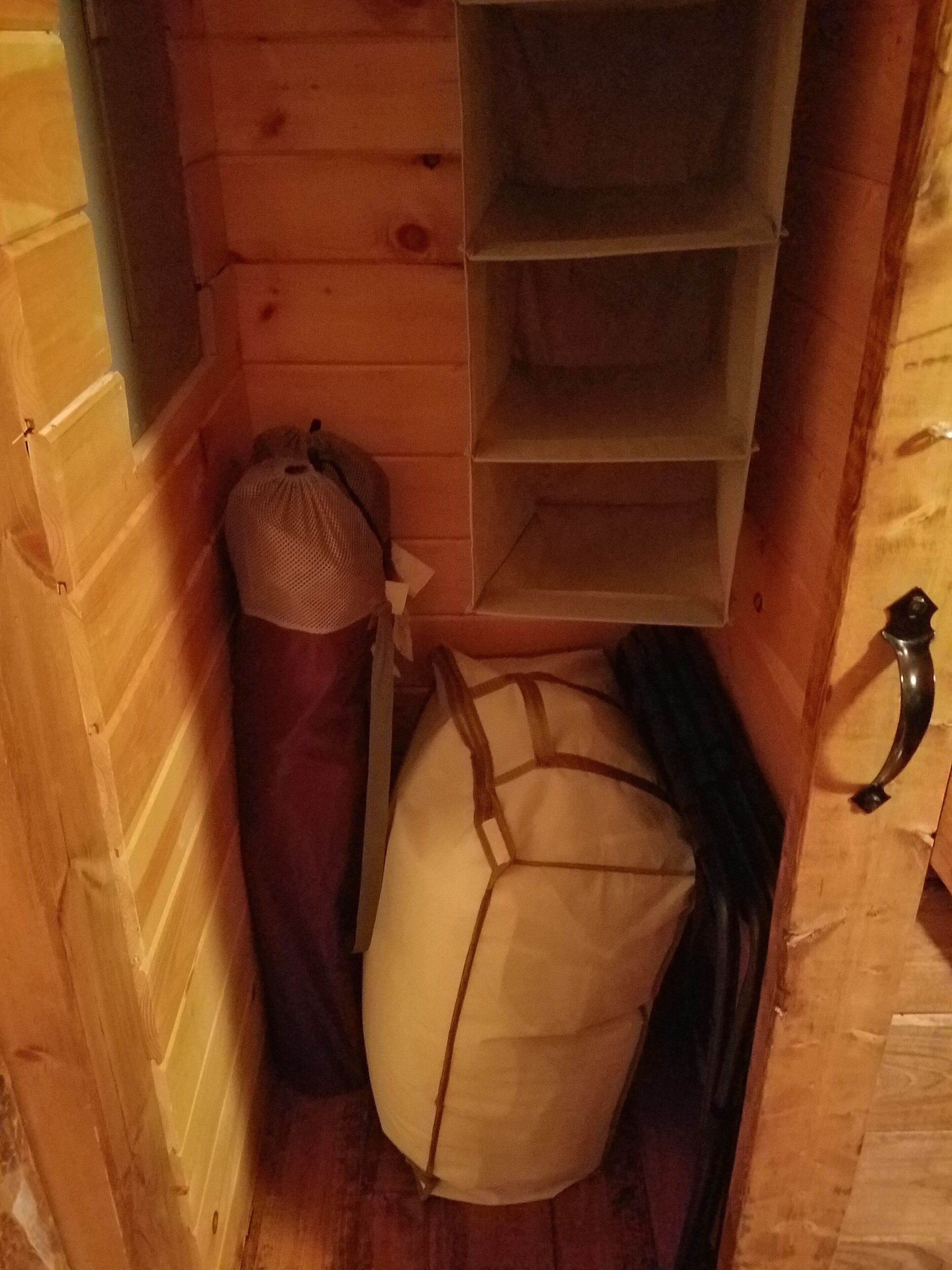 Une petite pièce étroite aux murs et au plancher en bois. Dans un coin, on trouve un grand objet blanc et rembourré, un petit sac et une poignée métallique sur le mur de droite.