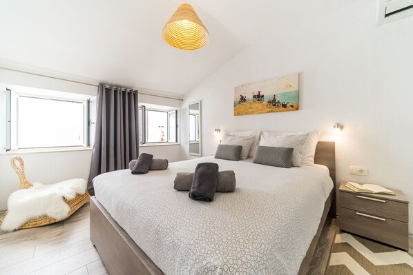 A bedroom with a large bed, a bedside table, a lamp, a painting on the wall, and a window with curtains.