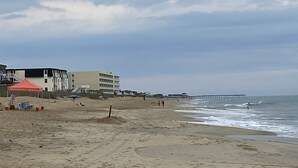 En strand med en mole, der strækker sig ud i vandet, bygninger i baggrunden og et rødt telt på sandet.
