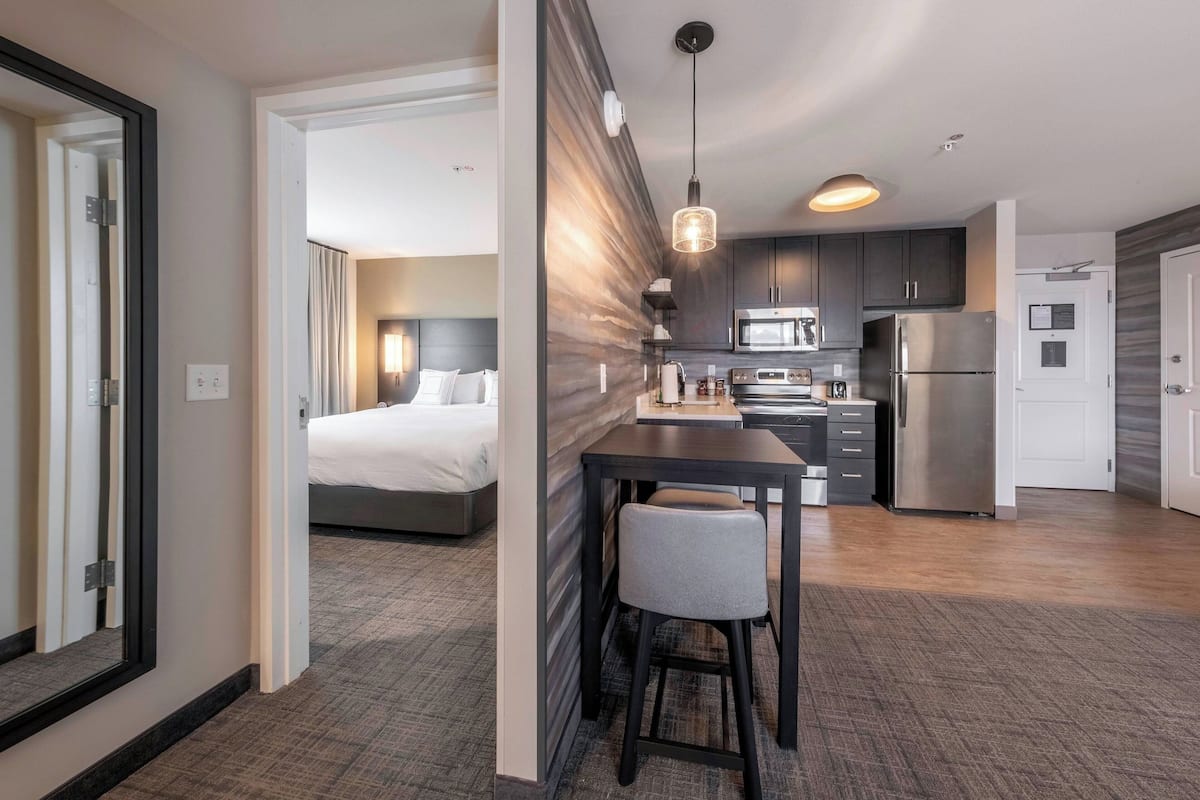 A modern hotel room with a bed, a small kitchen area with a microwave and refrigerator, and a dining area with a table and chairs.