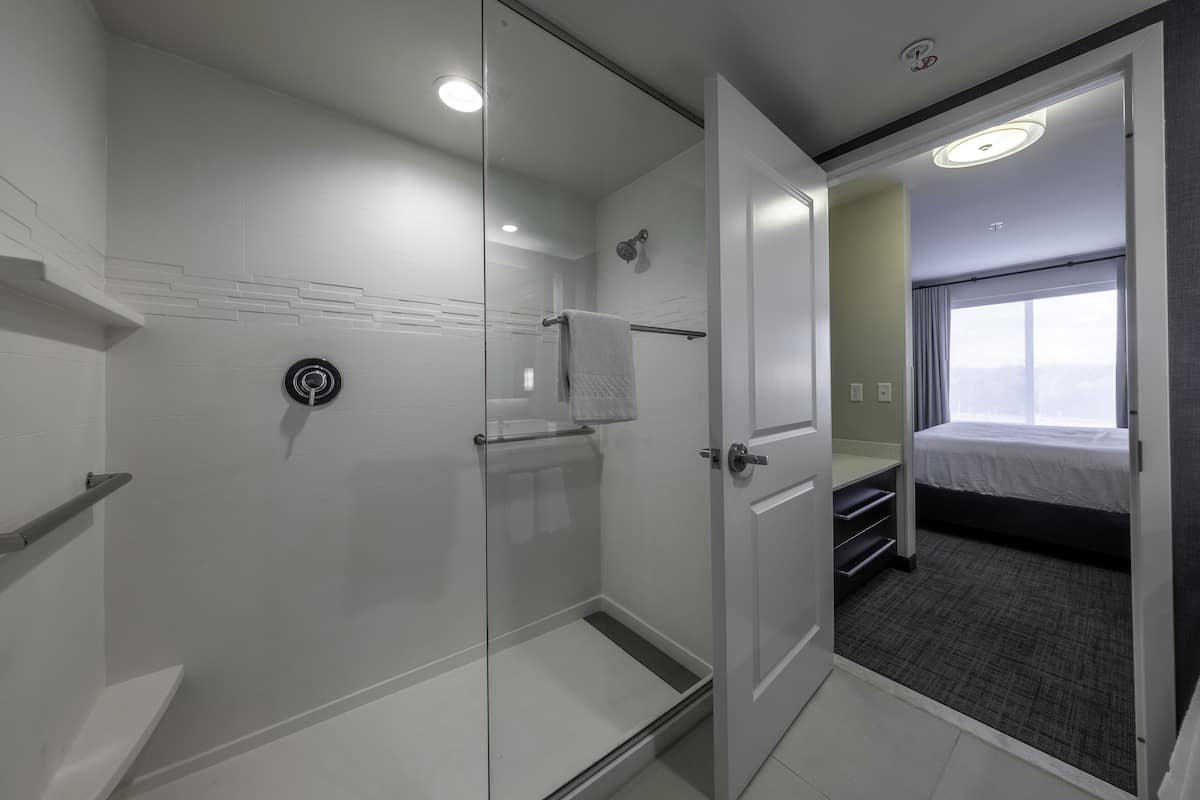 A modern hotel room with a glass shower enclosure, a bed with a gray mattress, and a view of the city through a large window.