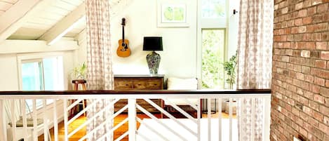 A spacious loft with a high ceiling, exposed wooden beams, a white staircase, and a balcony with a view of the greenery outside.