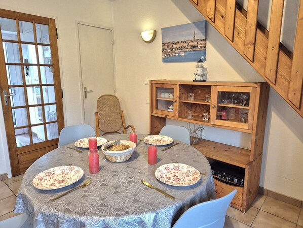 Ein Essbereich mit einem runden Tisch, gedeckt für vier Personen. Rund um den Tisch stehen Stühle. Eine Holzvitrine mit Glastüren befindet sich rechts, darin sind Gläser und Geschirr. Auf dem Tisch steht ein Korb mit Brot.