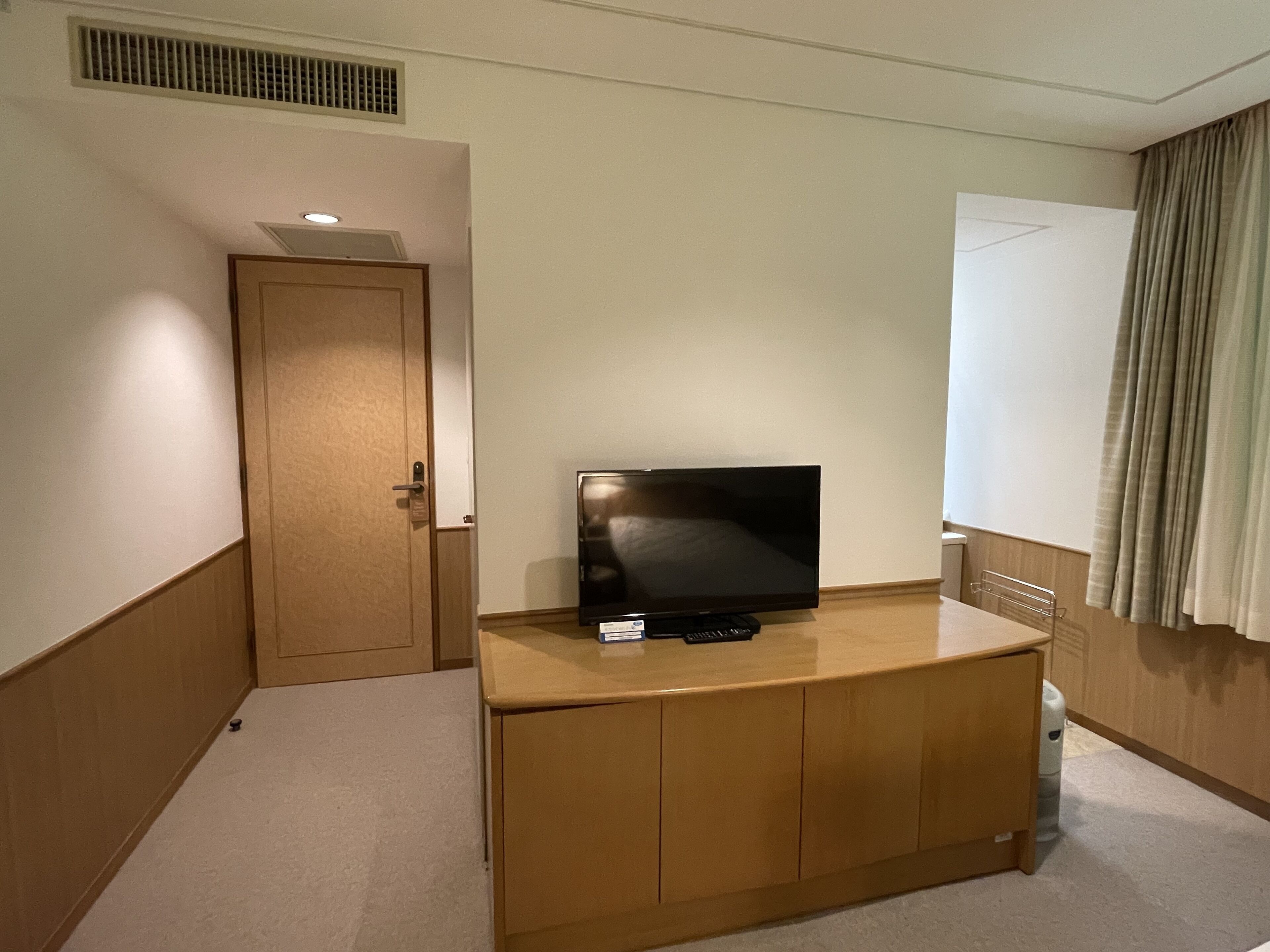 A hotel room with a wooden cabinet, a television, a door, and a window with curtains.