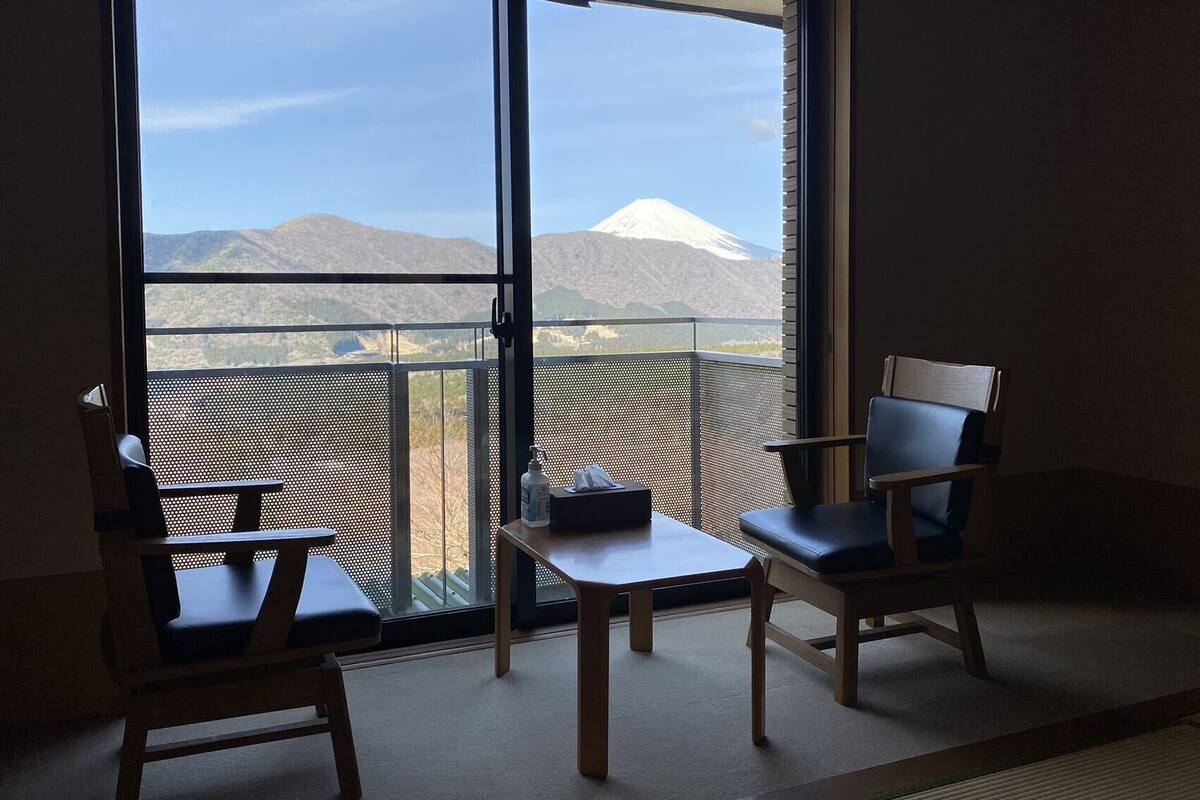 A room with two chairs and a small table, a sliding glass door leading to a balcony with a view of a mountain.