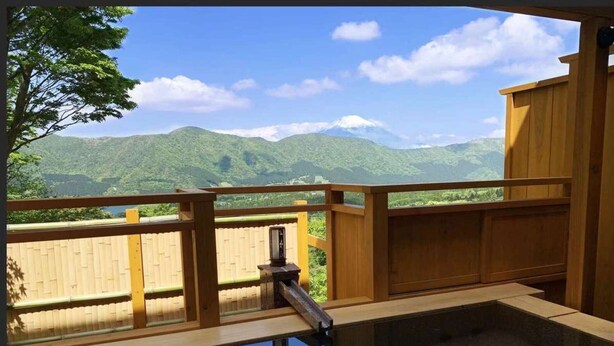Una terraza de madera con piscina de fondo de cristal, que ofrece vistas a montañas verdes y un cielo despejado.