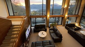 Amplia sala de estar con ventanas grandes que ofrecen vistas a montañas y un lago. Una escalera conduce a un nivel superior, y la habitación cuenta con muebles modernos y un televisor de pantalla plana.