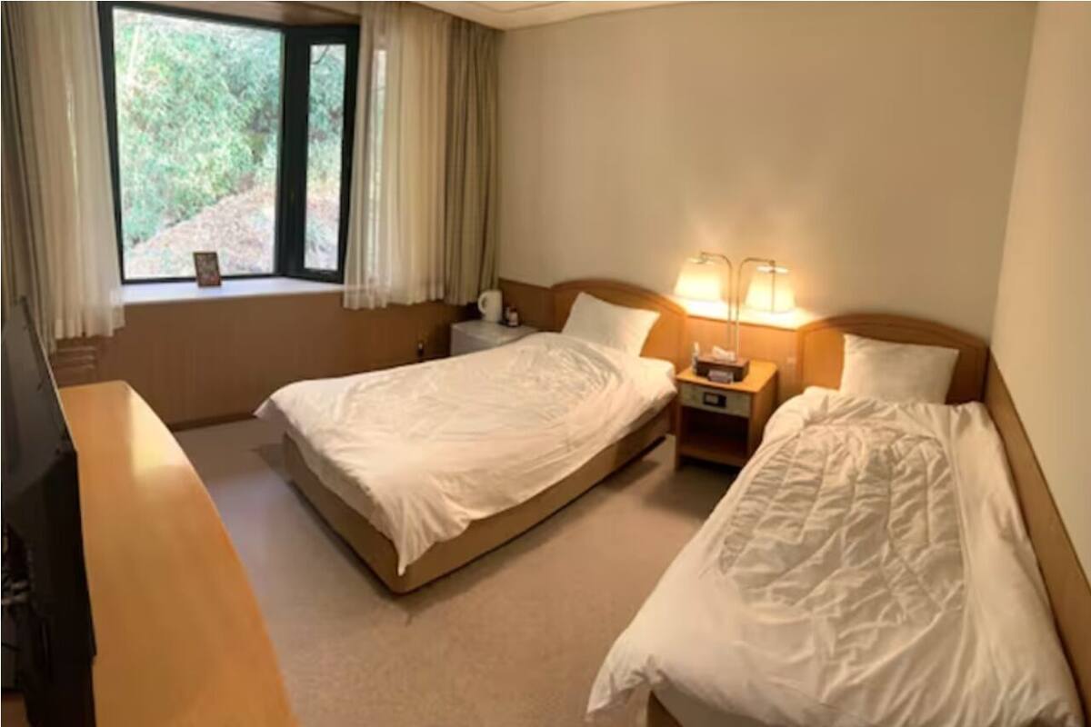 A hotel room with two single beds, a desk, a window with curtains, and a nightstand with a lamp.