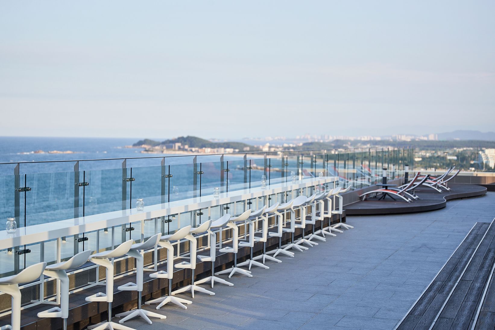 A seaside terrace with a glass railing, white chairs, and a view of the ocean and cityscape.