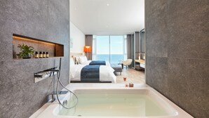 Habitación de hotel moderna con una bañera grande, una cama con dos almohadas, una mesita de noche y vistas al mar.