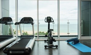 A gym with two treadmills and an elliptical machine, a blue yoga mat, and a view of the beach through large windows.