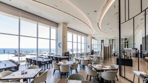 Un comedor amplio con mesas y sillas, ventanas grandes con vista al océano y una cocina moderna visible al fondo.