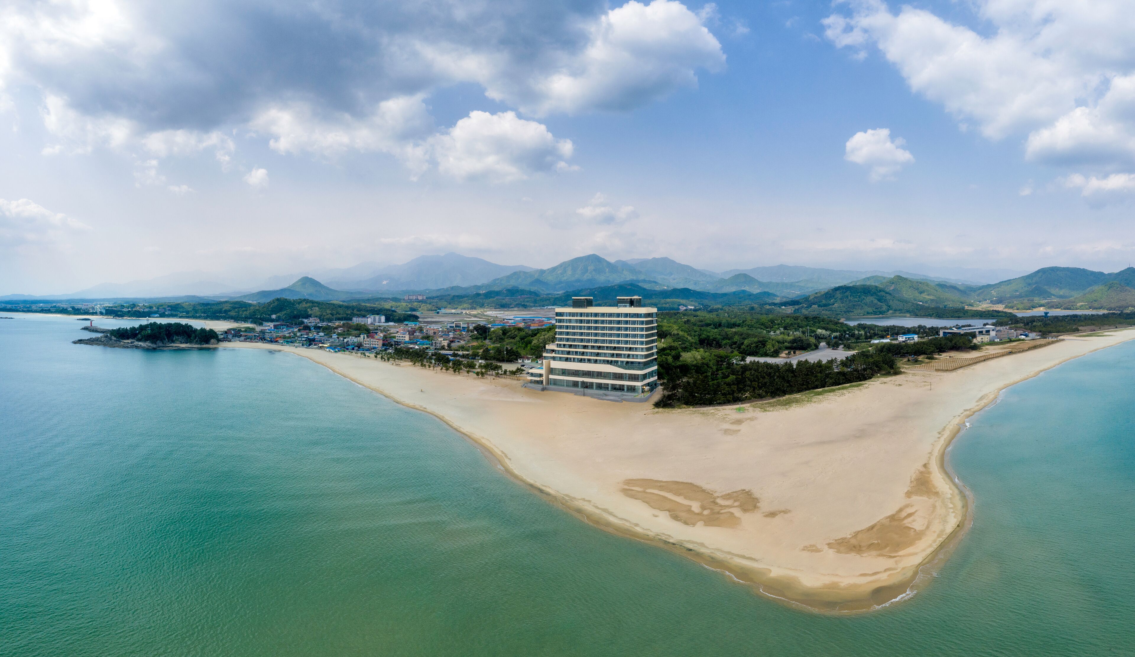 An aerial view of a coastal area with a prominent hotel building, a sandy beach, and a clear view of the surrounding mountains and water.