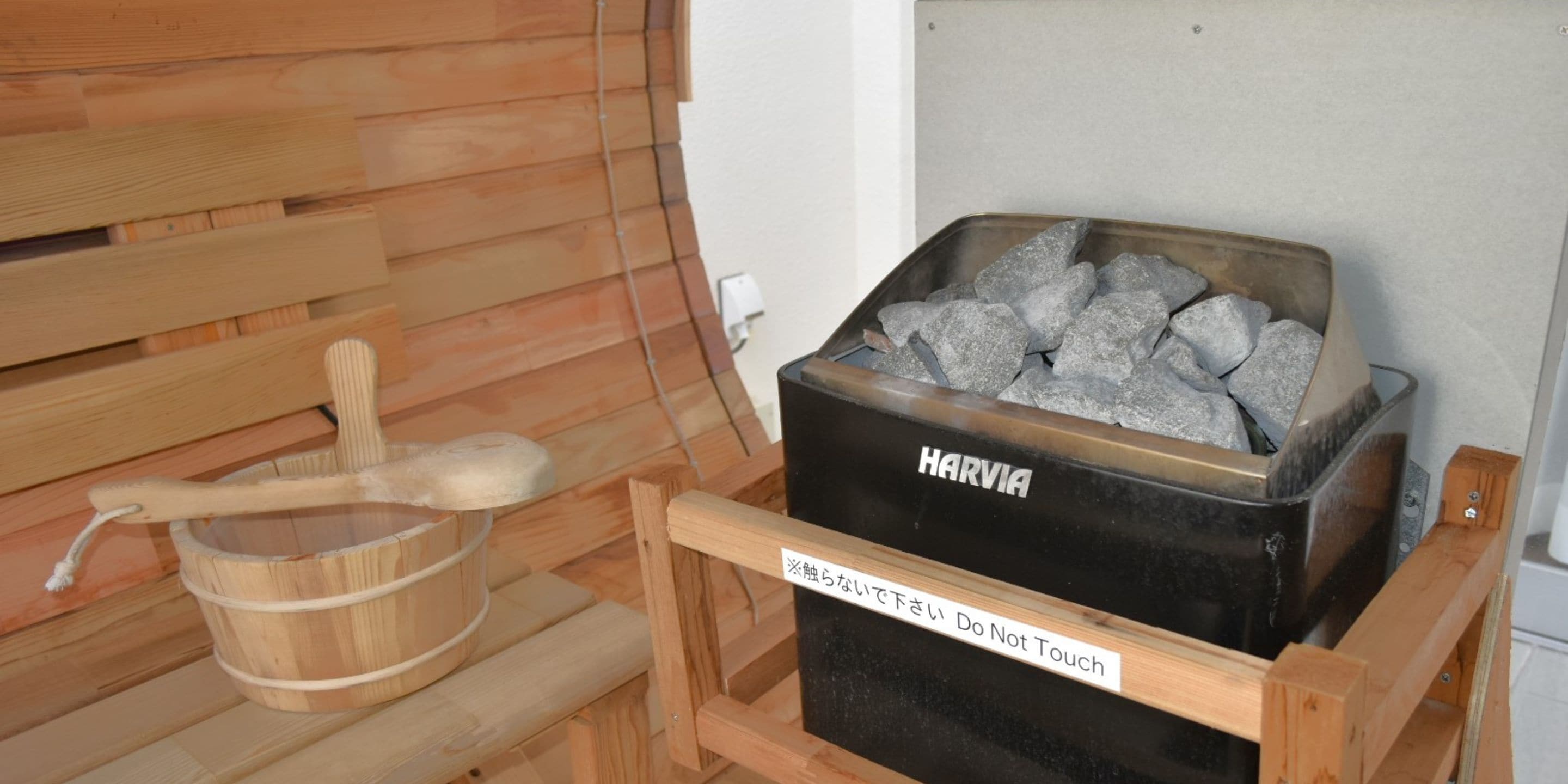 A sauna with a wooden bench, a wooden bucket, and a black container labeled "HARVIA" with a warning sign.