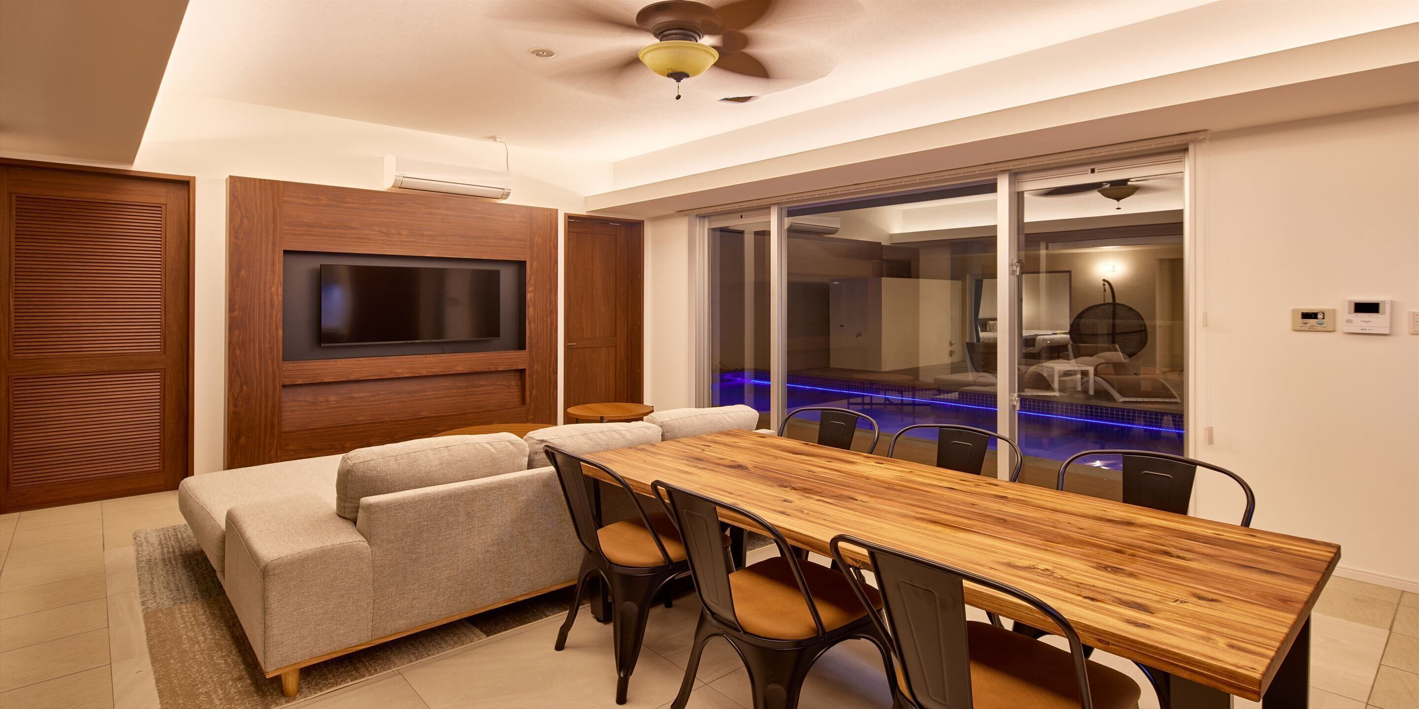 Una sala de estar moderna con una mesa de comedor de madera, asientos cómodos, un televisor de pantalla plana montado en la pared y una puerta de cristal que da a una zona de piscina.