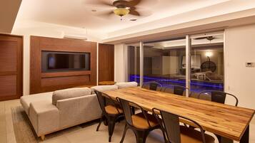 Una sala de estar moderna con una mesa de comedor de madera, asientos cómodos, un televisor de pantalla plana montado en la pared y una puerta de cristal que da a una zona de piscina.