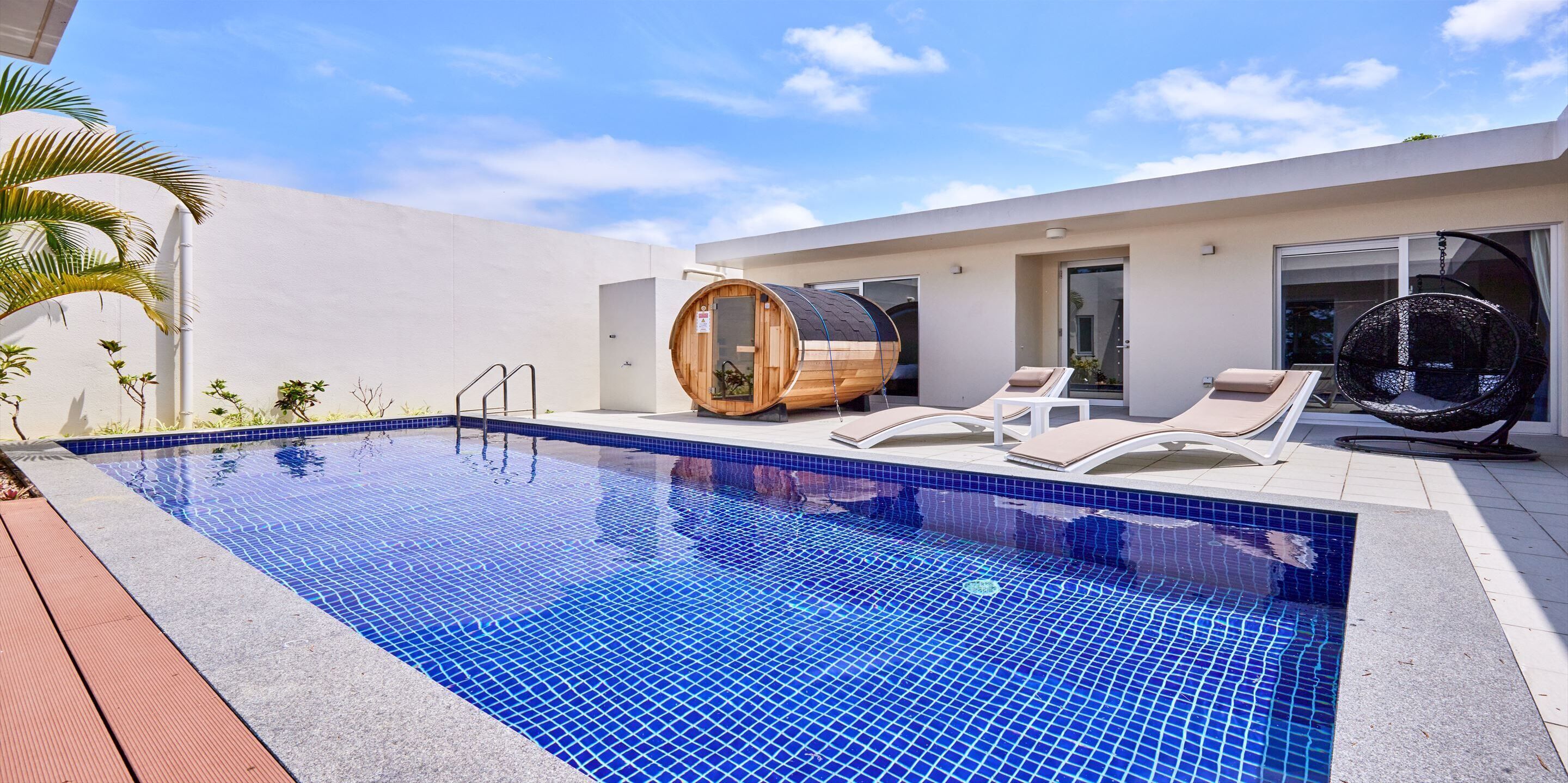 A modern outdoor swimming pool with a wooden sauna, lounge chairs, and a hanging chair.