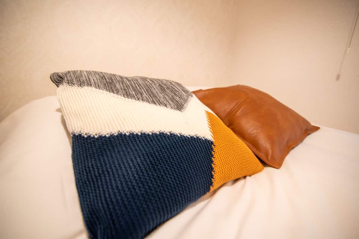 A bed with a white sheet, a blue and white knitted pillow, and a brown leather pillow.