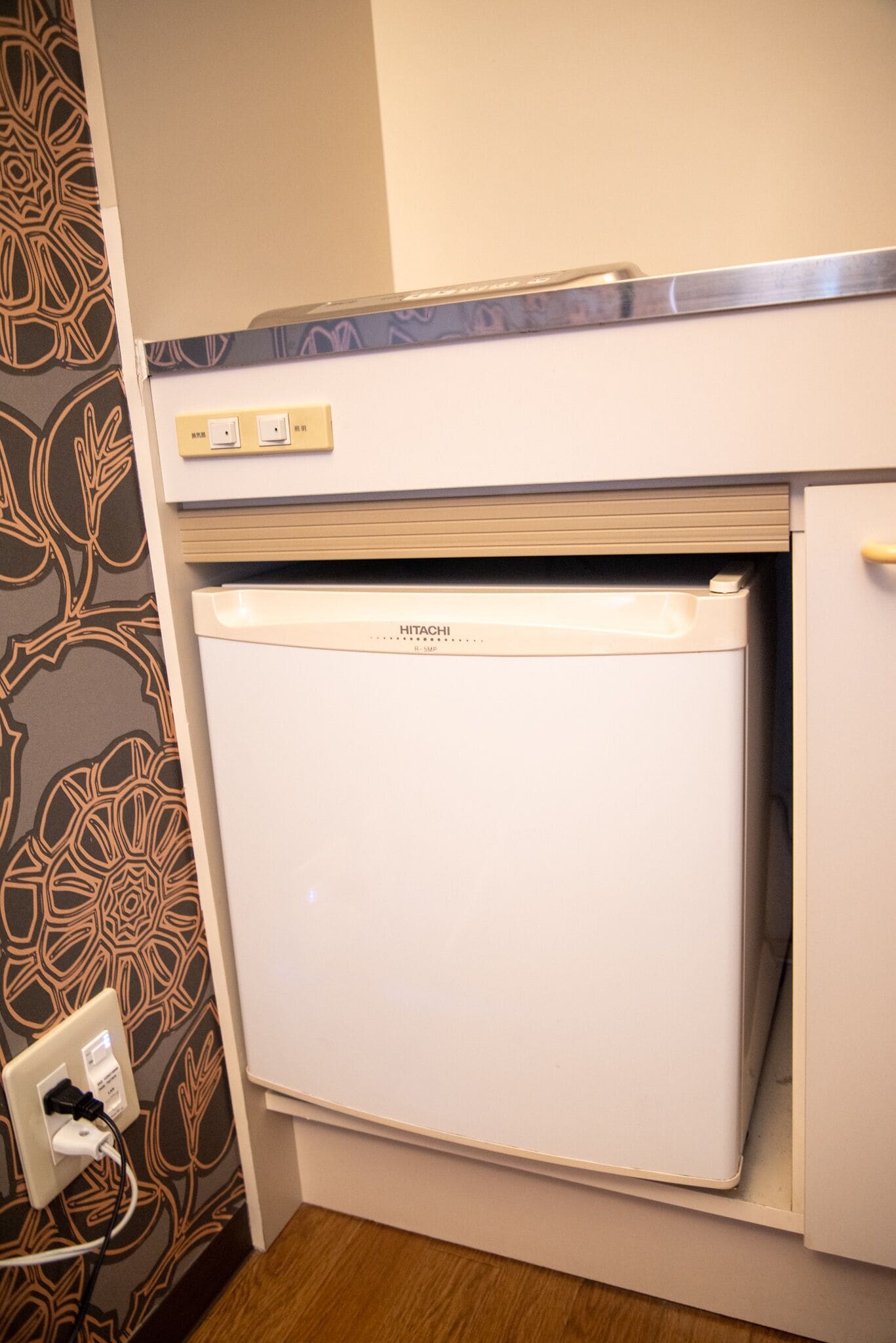 A compact refrigerator with a control panel and a freezer compartment, situated in a room with patterned wallpaper and a wooden floor.