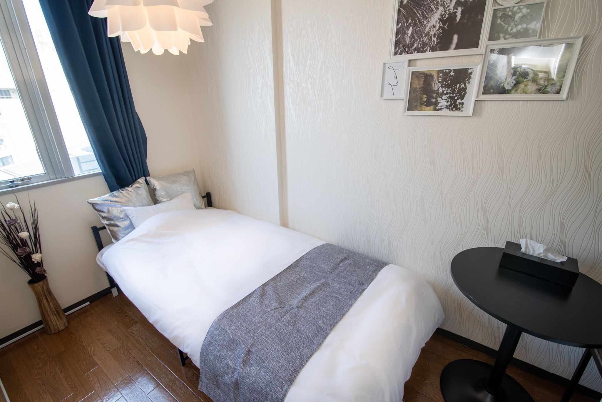 A neatly made bed with a gray throw blanket, a small bedside table with a tissue box, a vase with dried flowers, and a window with blue curtains.