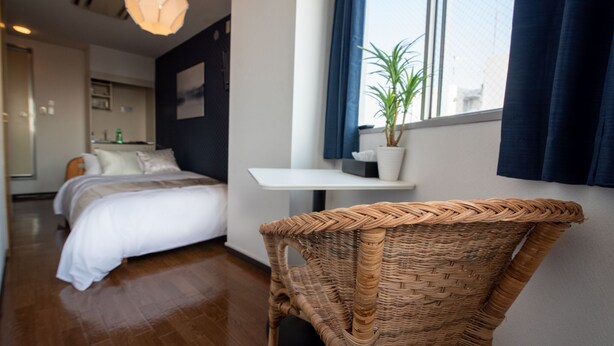 A hotel room with a bed, a wicker chair, a small table, a potted plant, and a window with curtains. - Vail sta. Takenotsuka (Tokyo)