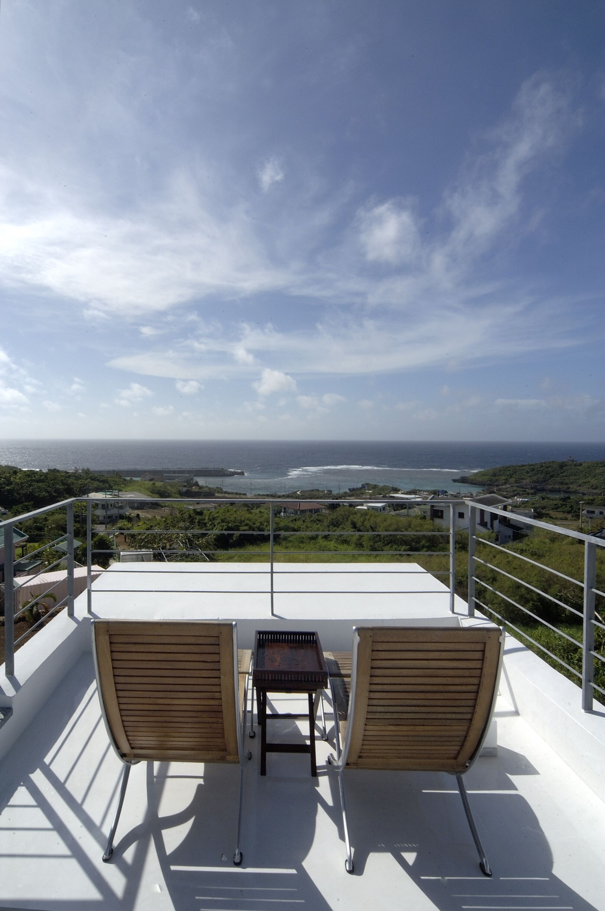 A balcony with two wooden chairs and a small table, overlooking a coastal landscape with buildings and greenery.