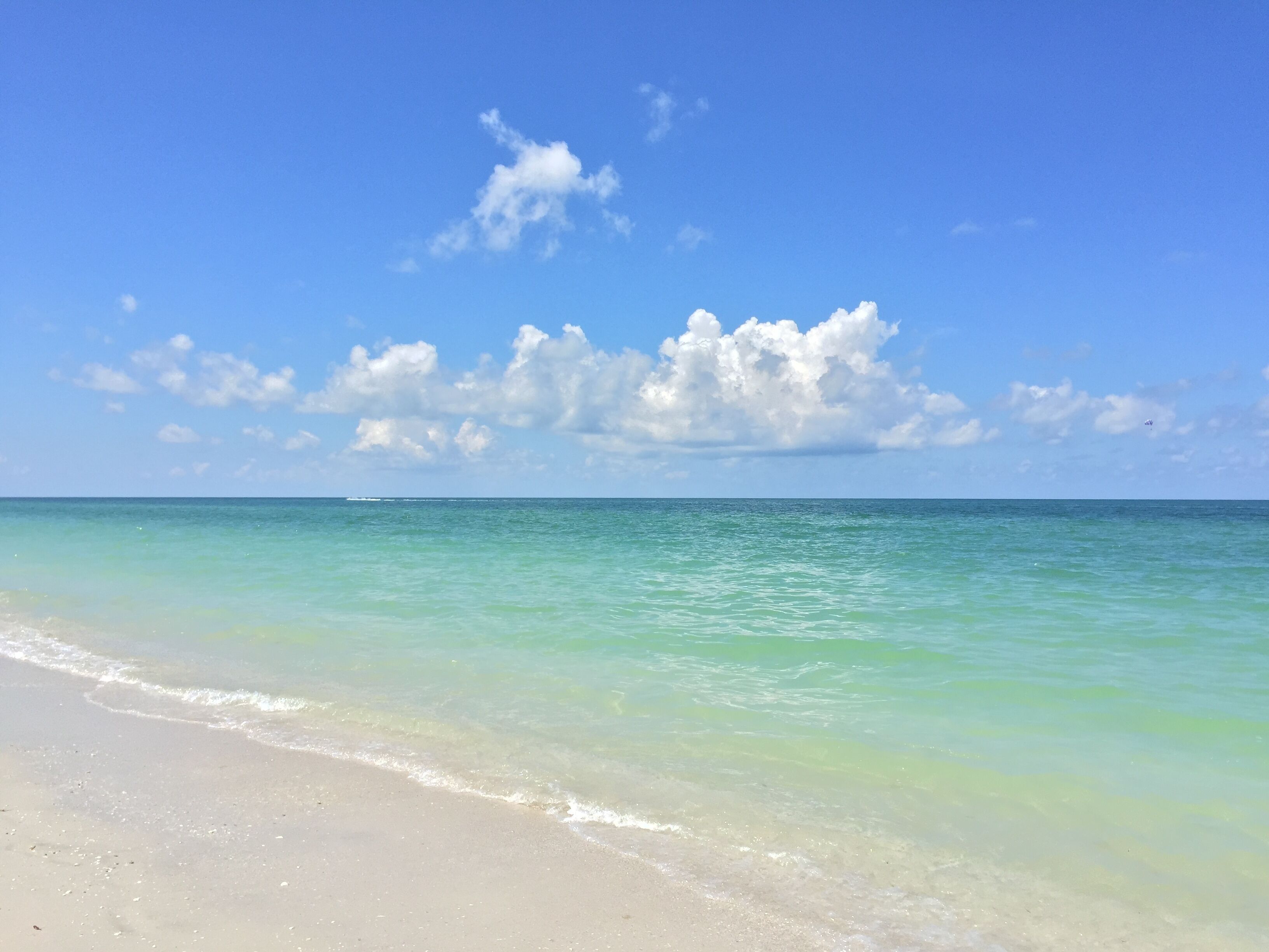 Une plage à l’eau turquoise et limpide, bordée d’un rivage sablonneux, sous un ciel bleu parsemé de quelques nuages.
