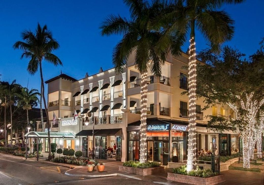 Een meerhoog hotel met balkons, palmbomen en winkelpanden.