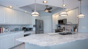 A modern kitchen with white cabinets, a marble island, stainless steel appliances, pendant lighting, and a ceiling fan. - The Dunes! Walk to the beach! Unwind in this amazing Naples retreat! (Naples)