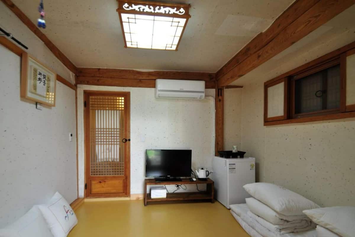 A room with a wooden ceiling, a television on a stand, a small refrigerator, and a futon bed.