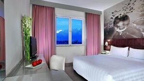 Una habitación de hotel con una cama grande, un escritorio con televisor, una silla y una ventana con cortinas rosadas.