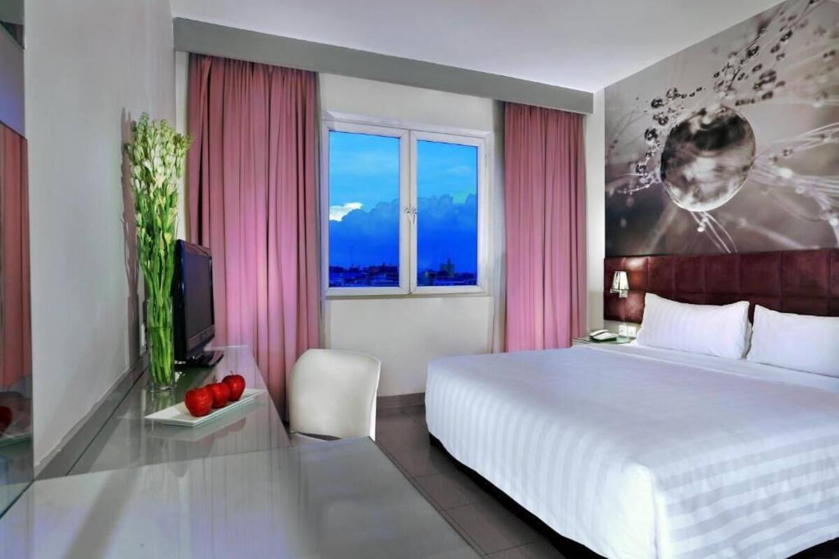 A hotel room with a large bed, a desk with a television, a chair, and a window with pink curtains.