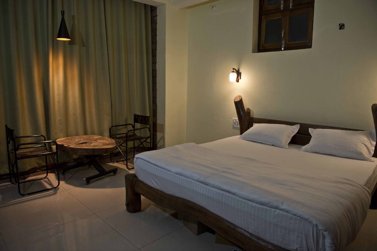 A bedroom with a wooden bed, a round table, two chairs, a hanging light, and a window with curtains.