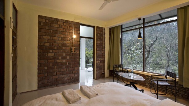 A bedroom with a large bed, a dining table, and chairs, a brick wall, and a view through large windows. - Corbett Riviera (Lansdowne)