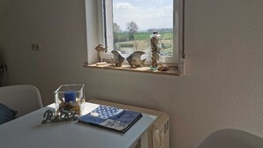Une pièce avec une fenêtre donnant sur un paysage rural, une table agrémentée d’un objet décoratif et une petite étagère avec des figurines.