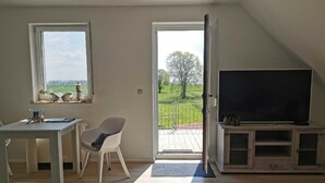Une pièce comprenant une table à manger, une chaise, un téléviseur posé sur un meuble en bois, et un balcon donnant sur un paysage verdoyant.