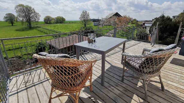 Une terrasse avec une table et des chaises en osier, donnant sur un paysage verdoyant avec des arbres et un bâtiment situé au loin.