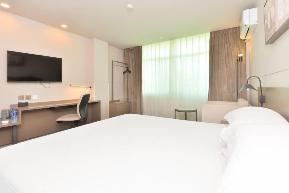 A hotel room with a bed, a desk with a chair, a TV, a window with curtains, and a bedside lamp.