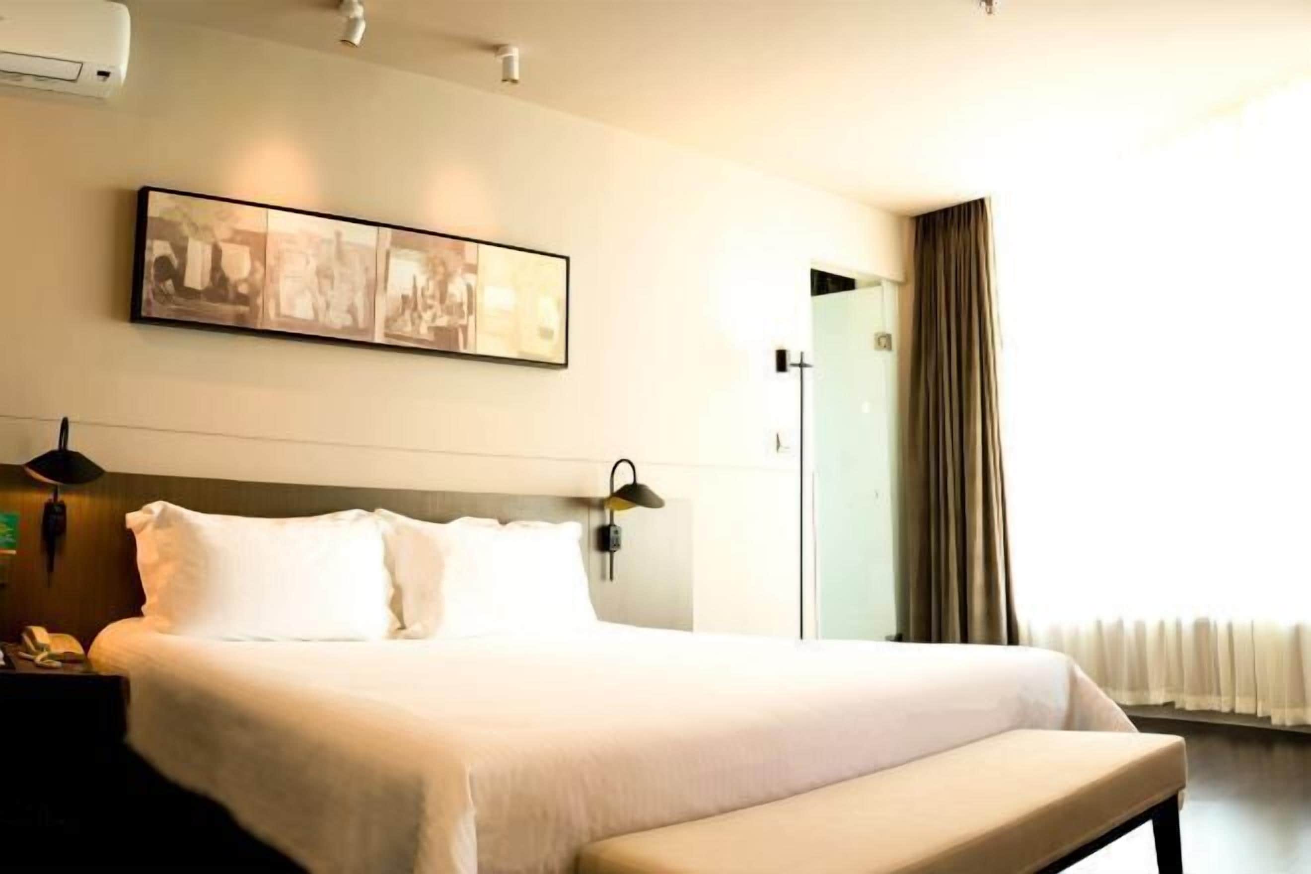 Quarto de hotel com uma cama grande, mesa de cabeceira com abajur, uma obra de arte na parede e uma janela com cortinas.