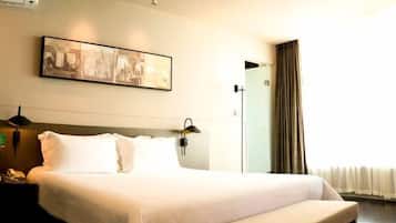 Quarto de hotel com uma cama grande, mesa de cabeceira com abajur, uma obra de arte na parede e uma janela com cortinas.