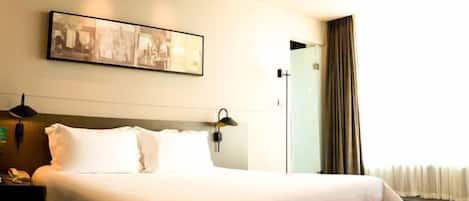 A hotel room with a large bed, a bedside table with a lamp, a wall-mounted artwork, and a window with curtains.