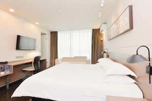 Ein Hotelzimmer mit einem großen Bett, einem Schreibtisch mit Stuhl, einem Flachbildfernseher, einer Schreibtischlampe und einem Fenster mit Vorhängen.
