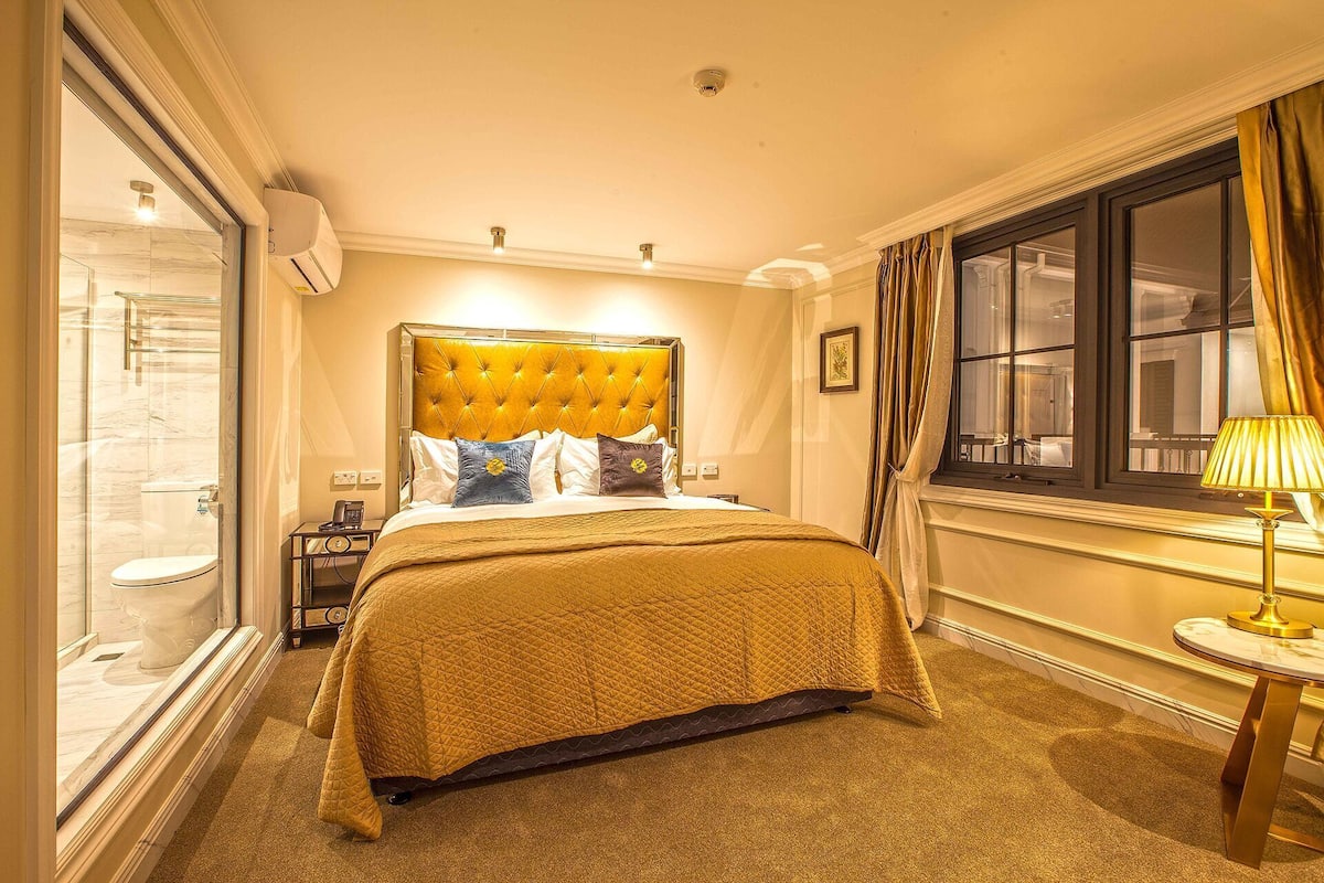 Une chambre d’hôtel avec un grand lit, une table de chevet, une fenêtre avec des rideaux et une lampe.