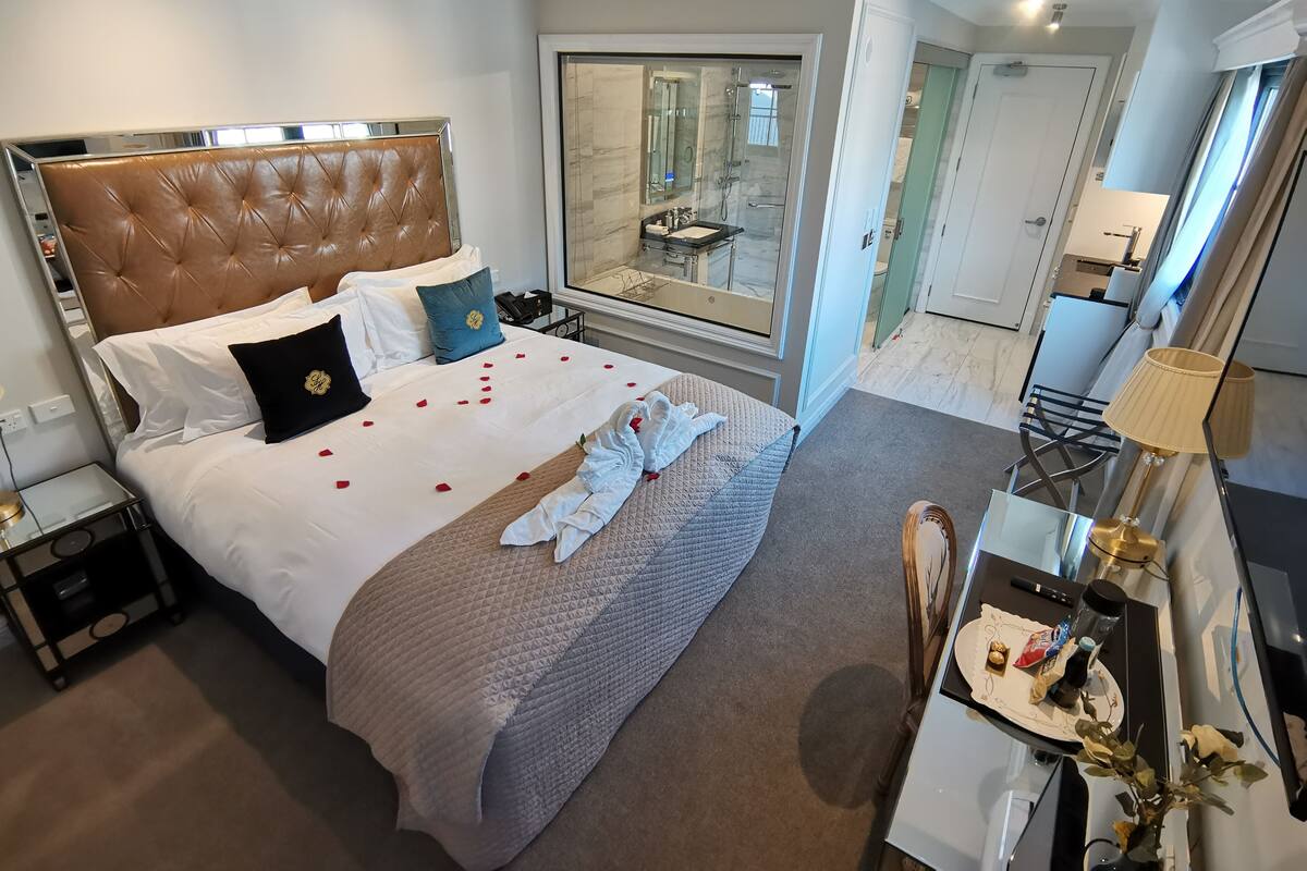 Une chambre d’hôtel avec un grand lit, une table de chevet, un bureau avec une lampe et une vue sur la salle de bain à travers une porte en verre.