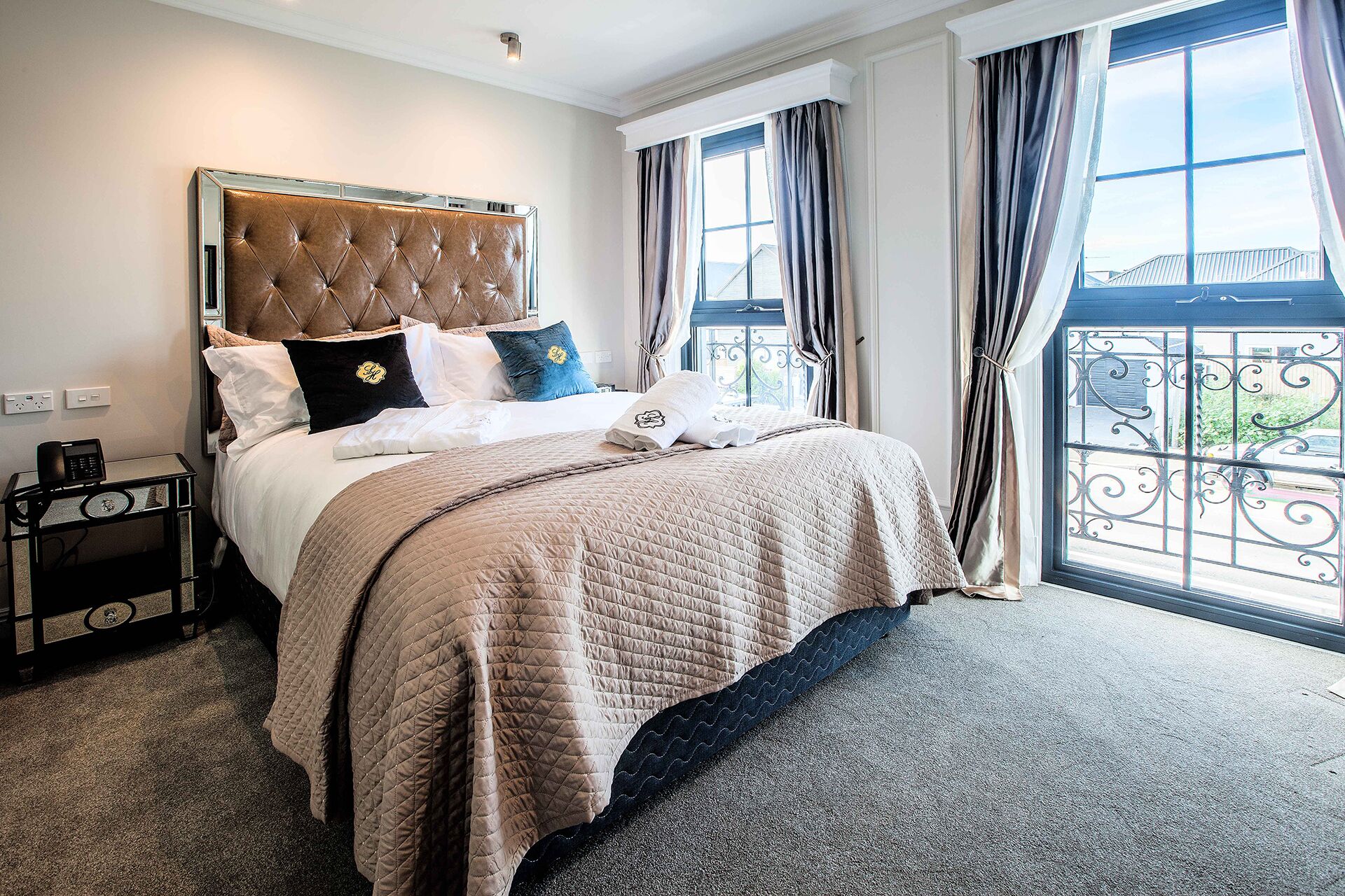 A hotel room with a large bed, a nightstand, a telephone, and a view of the outside through a window with curtains.