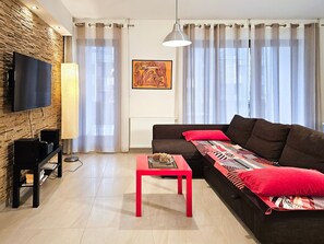 A modern living room with a flat-screen TV mounted on a textured wall, a red coffee table, a dark brown sofa with patterned cushions, and sheer curtains. - RS2.30 - Sienkiewicz Residence (RS1, RS2) (Swinoujscie)