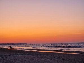 A beach scene with a person standing on the sand, waves approaching the shore, and a sunset in the background.