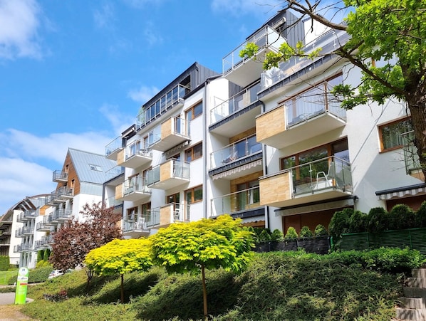 A multi-story residential building with balconies, a green park area, and a clear sky.
